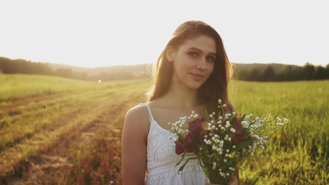 Pretty young woman in white dress standing in a meadow with bouquet of wild flowers on summer evening. Long hair flowing in wind in sunshine at sunset, she looks up into the camera and smiles