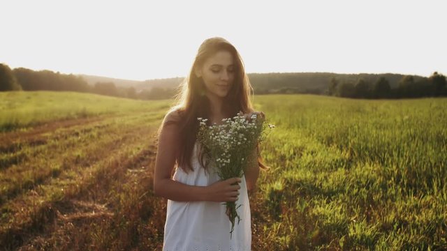 Pretty young woman in white dress standing in field with bouquet of wild flowers, loose long hair in the sun at sunset on a summer evening, looking at camera and smiling