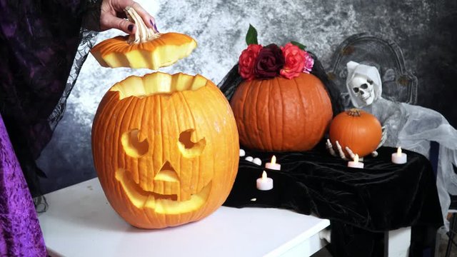 4k Woman puts candle inside Halloween Jack-o-lantern pumpkin on the table decorated with pumpkins, tombstone, ghost, candles and black velvet.