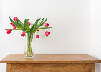 Red tulips with green leaves in glass vase on oak sidetable against white wall with copy space  (selective focus)