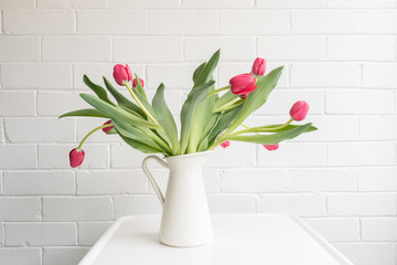 Red tulips with green leaves in white jug on table against painted brick wall