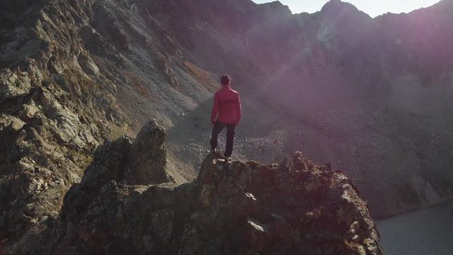A young hipster guy with a beard a climber in a cap and sunglasses is standing on a rocky ridge high in the mountains and looking around. Parkour in the mountains. Airview view. Movement around