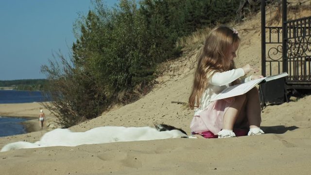 A sweet girl sits on the sand and reads a book strokes a dog