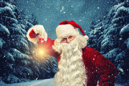 Santa Claus holds a lantern in his hands and looks into the camera against the background of a winter forest.