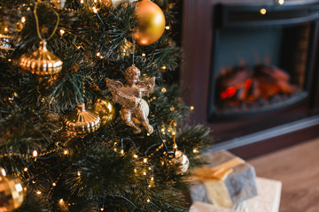 Cute toy golden angel hanging on decorated Christmas tree with gifts in cozy home