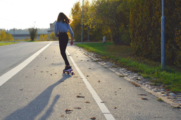 Teenage girl ride away skateboard or penny board on street back view against sunlight with shadow in autumn day.