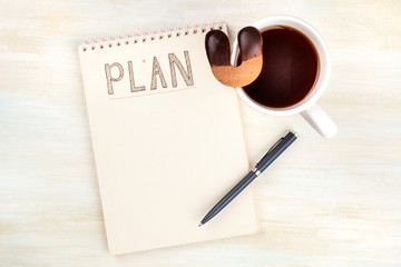 An overhead photo of a spiral notebook with the word Plan, with a cup of coffee with a cookie, on a desk with a place for text