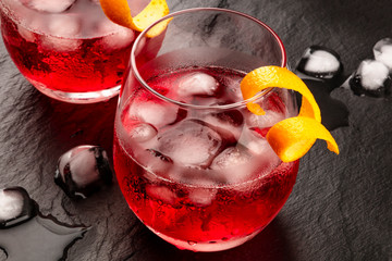 A photo of two vibrant cocktails with campari and orange twist garnishes, with ice cubes on a black...