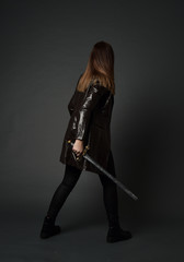 full length portrait of brunette girl wearing long leather coat and boots. standing pose and holding a sword on grey studio background.