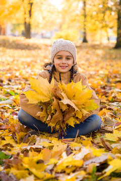 Autumn portrait: Cute smiling little girl sitting on the fallen leaves in autumn park and holding the bouquet of yellow maple leaves in her hands, vertical 
