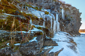 Fototapeta na wymiar The rocky cliff on the riverbank with moss and hanging icicles