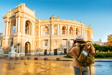 Obraz premium Woman traveler with dog in the backpack taking pictures with a camera while traveling. Odessa Opera Theatre. 