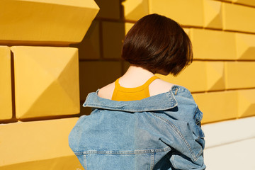 A brunette in a jeans jacket and yellow top is standing with her back against a yellow background.