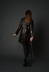 full length portrait of brunette girl wearing long leather coat and boots. standing pose and holding a sword on grey studio background.