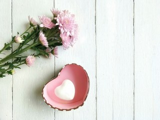 White ceramic heart shaped in pink gift box with pink chrysanthemum bouquet on white wooden table floor in love and valentine concept, top view with copy space