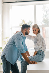 side view of smiling young couple hugging in kitchen while girlfriend sitting on kitchen counter
