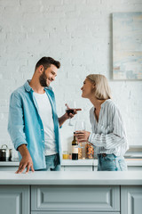 Fototapeta premium smiling young couple talking, holding glasses of red wine and looking at each other in kitchen