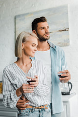 young couple hugging, holding glasses of red wine and looking away in kitchen
