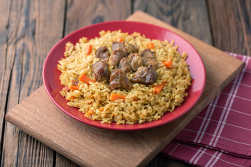 Red plate with pilaf on a brown wooden table. On the table is red pepper, green onions, garlic, cherry tomatoes, red napkin, spoon.