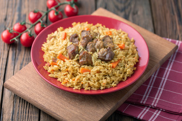 Red plate with pilaf on a brown wooden table. On the table is red pepper, green onions, garlic, cherry tomatoes, red napkin, spoon.