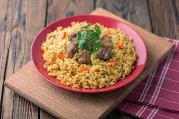 Red plate with pilaf on a brown wooden table. On the table is red pepper, green onions, garlic, cherry tomatoes, red napkin, spoon.