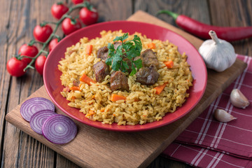Red plate with pilaf on a brown wooden table. On the table is red pepper, green onions, garlic, cherry tomatoes, red napkin, spoon.