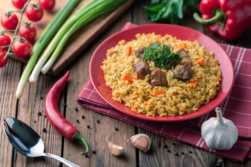 Red plate with pilaf on a brown wooden table. On the table is red pepper, green onions, garlic, cherry tomatoes, red napkin, spoon.