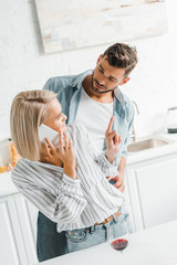 young girlfriend talking by smartphone and showing one finger up to boyfriend in kitchen