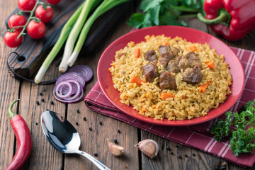 Red plate with pilaf on a brown wooden table. On the table is red pepper, green onions, garlic, cherry tomatoes, red napkin, spoon.