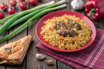 Red plate with pilaf on a brown wooden table. On the table is red pepper, green onions, garlic, cherry tomatoes, red napkin, spoon.