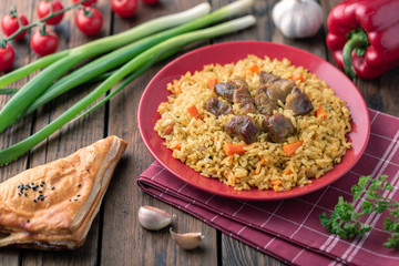 Red plate with pilaf on a brown wooden table. On the table is red pepper, green onions, garlic, cherry tomatoes, red napkin, spoon.