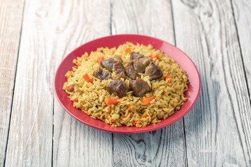 Red plate with pilaf on a white wooden table. On the table is red pepper, green onions, garlic, cherry tomatoes, white napkin, spoon.