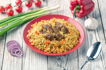 Red plate with pilaf on a white wooden table. On the table is red pepper, green onions, garlic, cherry tomatoes, white napkin, spoon.