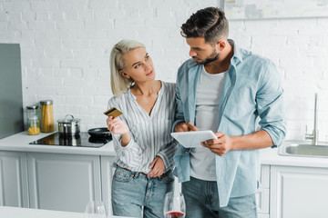 boyfriend holding tablet and girlfriend standing with credit card in kitchen