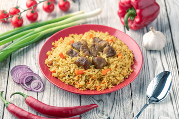 Red plate with pilaf on a white wooden table. On the table is red pepper, green onions, garlic, cherry tomatoes, white napkin, spoon.