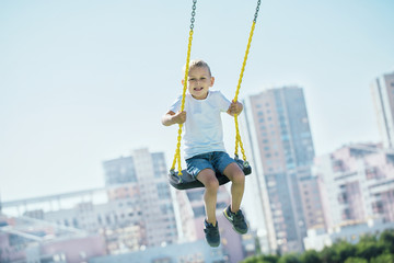Little boy looks into the camera and enjoys the swings on the playground in the city park