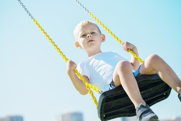 Little boy looks into the camera and enjoys the swings on the playground in the city park