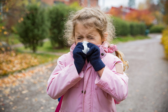 Sick little girl with cold and flu standing outdoors. Preschooler sneezing, wiping nose with handkerchief, coughing, having runny red nose. Autumn street background