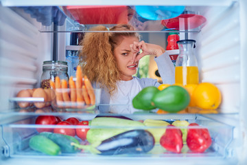 Womann standing in front of opened fridge and holding up to her nose because of bad smell. Picture...