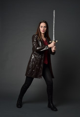 full length portrait of brunette girl wearing long leather coat and boots. standing pose and holding a sword on grey studio background.