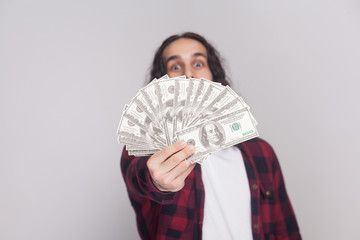 Portrait of cunning young businessman in red checkered shirt and long curly hair standing, keeking and demonstrating fan of money and looking at camera. Indoor studio shot, isolated on gray background