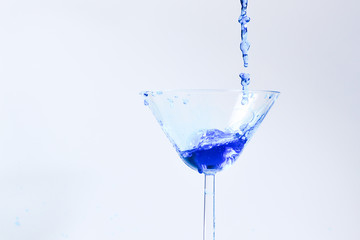 Cocktail with blue liquid in glass. Glass with blue water pouring with liquid with splashes and drops. Martini glass filling with alcohol with splashes on white background. Refreshing drink concept.