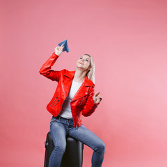 Happy young woman with travel suitcase launches paper airplane. Blonde tourist girl on pink background, concept