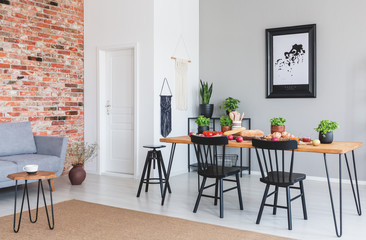 Black chairs at dining table and poster in flat interior with grey sofa against red brick wall. Real photo