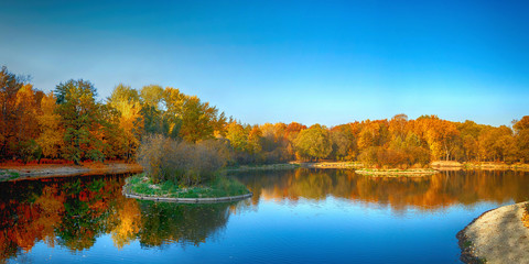 Panorama of a beautiful golden autumn forest with a lake in sunny weather with bright blue sky