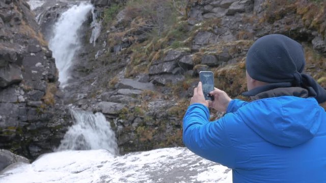 A man photographs a beautiful waterfall in the mountains, on a smartphone. Leads an active and interesting life and puts photos from their travels on social networks.