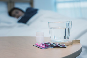 Closeup on pills and glass of water on table and Asian women sleep sick on bed in bed room.color tone