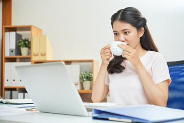 Pretty Asian woman having cup of coffee while watching laptop at table in new office