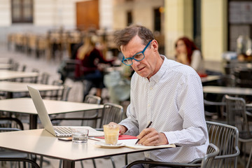 Happy attractive stylish mature man working on laptop while having coffee in outside coffee shop