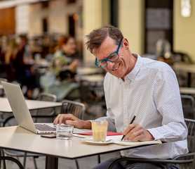 Happy attractive stylish mature man working on laptop while having coffee in outside coffee shop